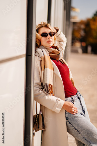 student or businesswoman in sunglasses and a coat with scarf walks down a street © andrey
