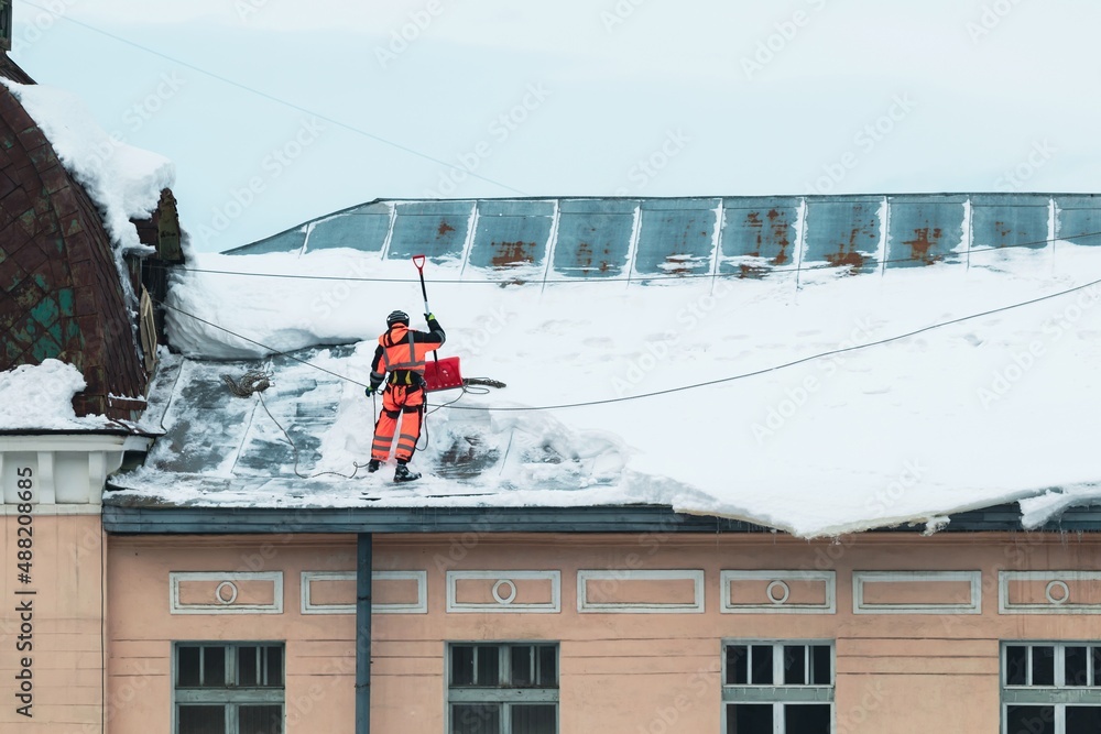 Obraz premium A working man in bright overalls with a safety belt with a shovel clears snow from the roof of an old building. Prevention of snow falling from the roof, industrial mountaineering