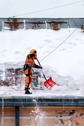 A working man in bright overalls with a safety belt with a shovel clears snow from the roof of an old building. Prevention of snow falling from the roof, industrial mountaineering