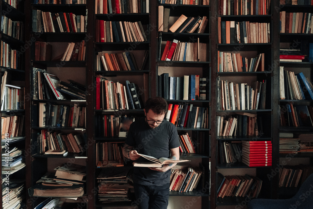 Adult student reading a book in the college library. Young standing male wearing glasses with bookshelves on background.