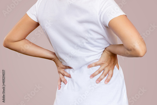 Cropped shot of a young woman in a white t-shirt holding her lower back with her hands isolated on color beige background. Back pain. Sciatica, osteochondrosis, gallstone disease, pancreatitis
