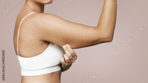 Close-up of a young tanned woman grabbing skin on her upper arm with excess fat isolated on a beige background. Pinching the loose and saggy muscles. Overweight concept photo