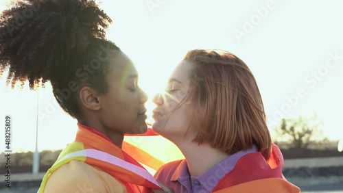 Mulitracial lesbian girls kissing together at gay pride parade while holding rainbow flag - LGBT concept photo