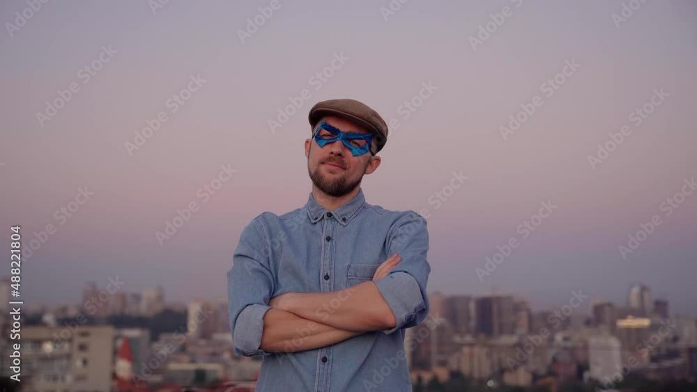 Cute bearded male in blue superhero mask and jeans shirt posing in front of the camera putting on peaked cap outdoor at sunset with urban view and crossing arms. Power or justice concept. 4k footage Cute bearded male in blue superhero mask and jeans shirt posing in front of the camera putting on peaked cap outdoor at sunset with urban view and crossing arms. Power or justice concept. 4k footage