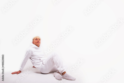 Attractive sporty young man with blond hair wearing white sportswear sitting on white background, looking up