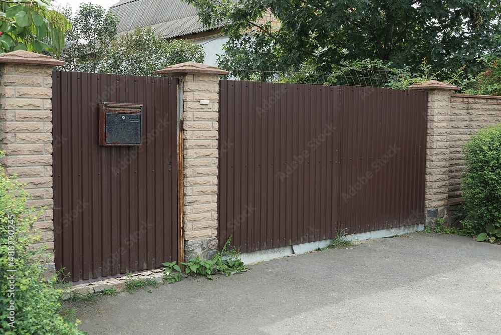 Fototapeta premium one big closed brown metal gate and part of the wall of the fence made of bricks in green vegetation on the street
