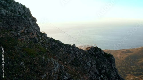 Aerial shot tracking forward over the edge of Table Mountain to dramatically reveal the shore of Camps Bay Cape Town during a beautiful golden sunset.