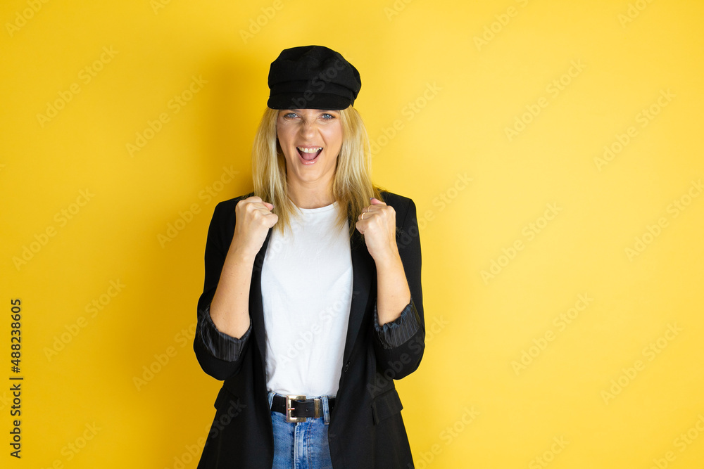 Beautiful woman wearing casual white t-shirt and a cap over isolated yellow background very happy and excited making winner gesture with raised arms, smiling and screaming for success. Beautiful woman wearing casual white t-shirt and a cap over isolated yellow background very happy and excited making winner gesture with raised arms, smiling and screaming for success.