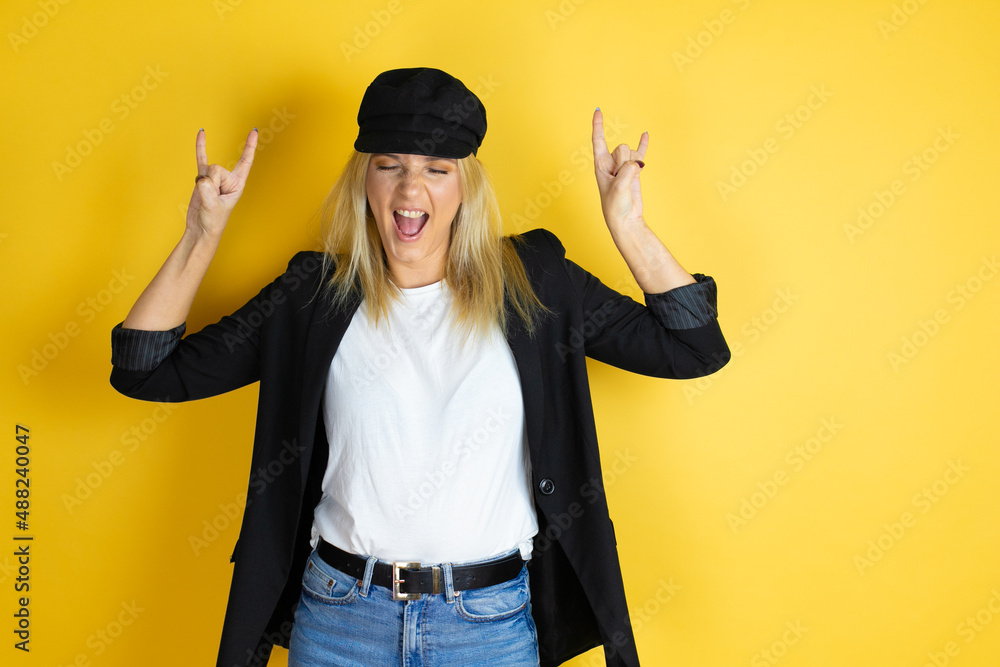 Beautiful woman wearing casual white t-shirt and a cap over isolated yellow background shouting with crazy expression doing rock symbol with hands up Beautiful woman wearing casual white t-shirt and a cap over isolated yellow background shouting with crazy expression doing rock symbol with hands up