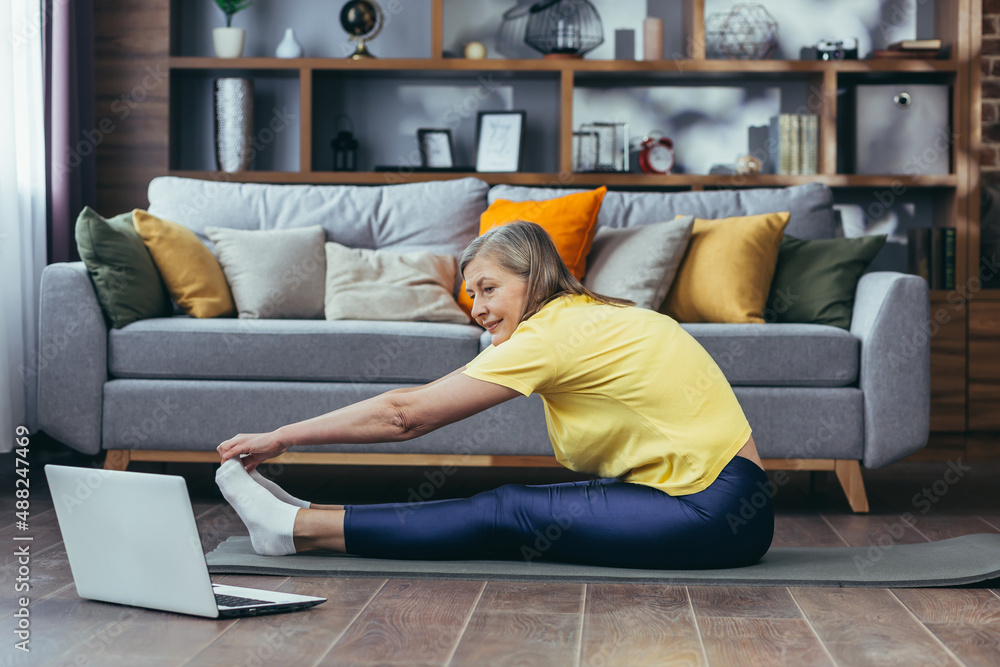 Obraz premium Senior gray-haired woman doing fitness at home sitting on a mat, online aerobics classes, doing stretching, looking at laptop screen