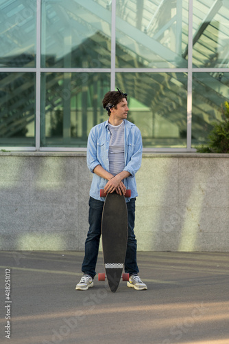 Street style man holding longboard over modern city building. Hipster guy skateboarder with skateboard outdoors. Trendy young male hipster holding long board. Urban lifestyle and youth freedom concept