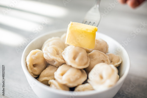 Closeup macro of traditional Russian dumplings pelmeni with meat filling stuffing in white bowl with hand fork placing butter on hot food in traditional recipe