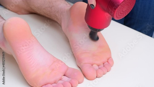 Hand of unrecognized female masseur with electric percussion vibratory massager massages the soles of the athlete's feet. Sports body regenerating massage therapy, close-up view photo
