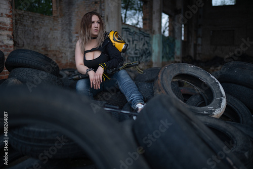 A beautiful strong girl with big breasts sits with a machine gun and a gas mask on her hip the ruins of a destroyed building and a pile of old tires. Guerrilla, rebel or resistance fighter