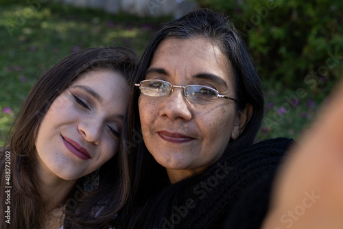 Latin American mother (50) and daughter (24) take a selfie in the park, the daughter lovingly leans on her mother's shoulder closing her eyes. Mother's day concept and technology.