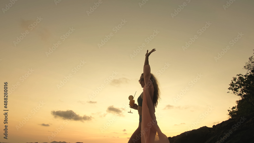 Silhouette woman stand on a reef rock stone in sea on golden sunset. Girl on tropical beach in green swimsuit and flutter in wind pink tunic silk shirt cape, drinks her orange cocktail Pina Colada