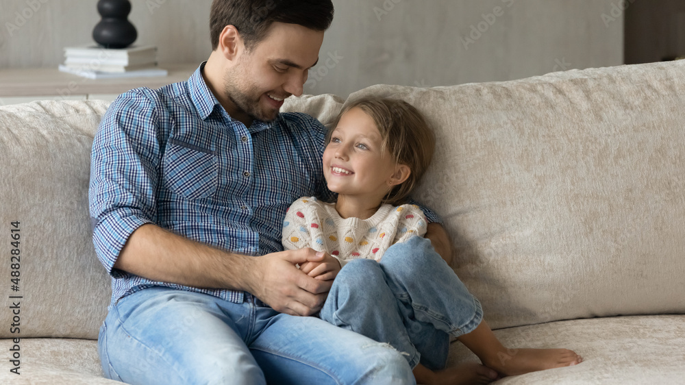 Happy loving dad cuddling, hugging little daughter on couch with tenderness, care, talking to child, laughing, enjoying family leisure time, friendship with child, being father, fatherhood Happy loving dad cuddling, hugging little daughter on couch with tenderness, care, talking to child, laughing, enjoying family leisure time, friendship with child, being father, fatherhood