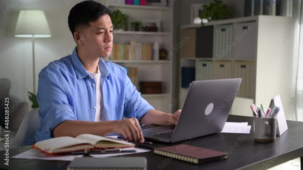 Young Asian businessman sitting at home working on laptop computer. Asian man studying online using laptop surfing information Young Asian businessman sitting at home working on laptop computer. Asian man studying online using laptop surfing information