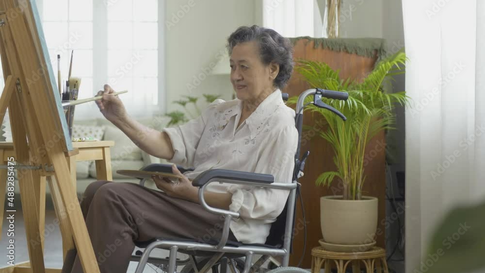 Senior woman painting picture at living room. She sitting on wheelchair while painting pictures. Senior woman painting picture at living room. She sitting on wheelchair while painting pictures.