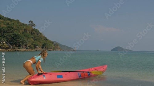 Slim young woman pushes pink plastic canoe to clear ocean water from sandy beach at exotic resort on sunny day. Traveling to tropical countries. Attractive sexy girl posing near kayak. © ivandanru