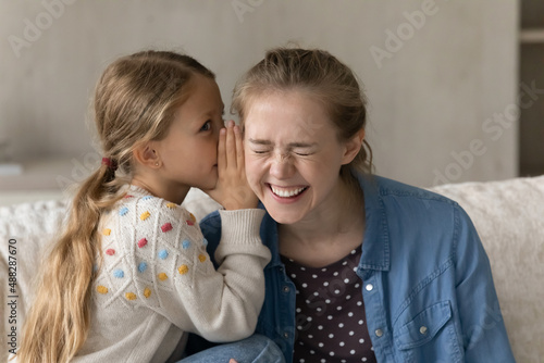 Cute kid telling secret to happy laughing elder sister, nanny, daycare teacher, whispering in ear. Pretty loving daughter girl and young mom enjoying friendship, having fun, talking at home photo
