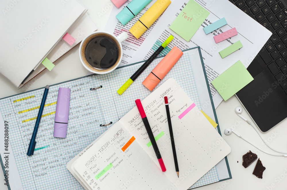Student's workspace. Top view of the table filled with notes, notebooks, stickers and markers and a cup of coffee. A notebook with a study schedule on the table. Student's workspace. Top view of the table filled with notes, notebooks, stickers and markers and a cup of coffee. A notebook with a study schedule on the table.