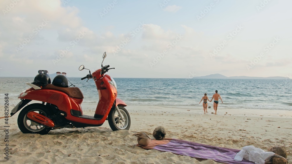 Foto Stock Scooter road trip. Lovely couple on red motorbike in white  clothes on sand beach. Just married people kiss hugs walking near the  tropical palm trees, sea. Wedding honeymoon by ocean.