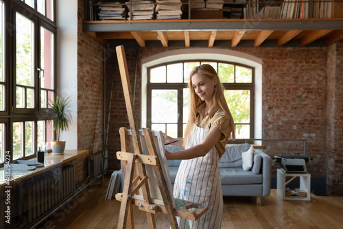Motivated inspired smiling millennial blonde woman in apron drawing pictures with paints on easel, standing in modern studio or living room. Happy skilled young female artist working on new artwork.