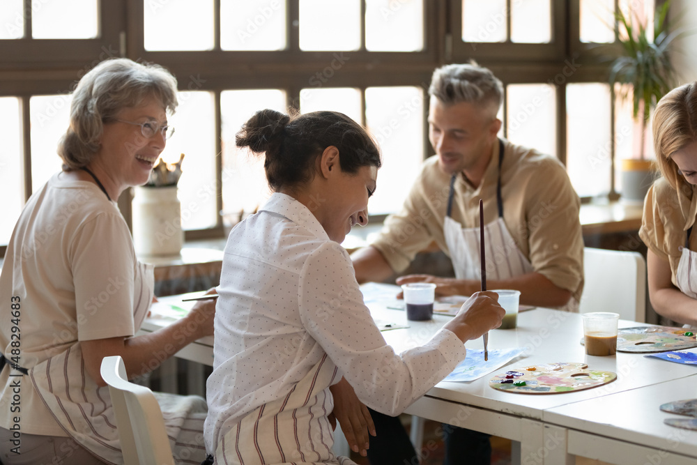 Side back view happy millennial Indian woman talking with diverse people, enjoying drawing pictures on creative art masterclass sitting together at table in modern loft stylish school, hobby activity.