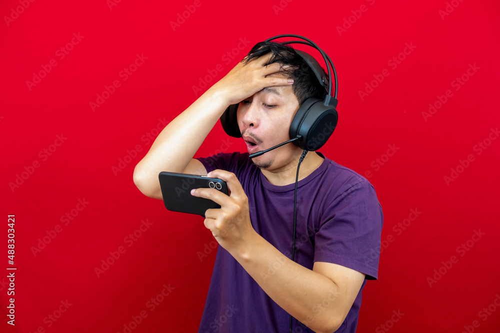 An asian man is playing a losing game on his smartphone and wearing a headset. An asian man is playing a losing game on his smartphone and wearing a headset.