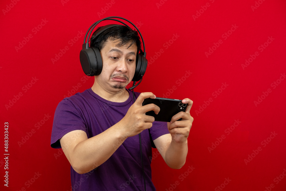 An asian man is playing a game on his smartphone and wearing a headset. An asian man is playing a game on his smartphone and wearing a headset.