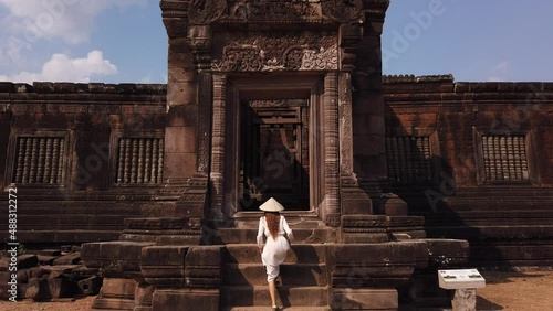 Beautiful woman in long white dress and vietnamese hat going up the stairs to palace in Wat Phou ruined Khmer Hindu Temple complex. Champassak, Laos, Asia. Ancient culture religious architecture. Slow