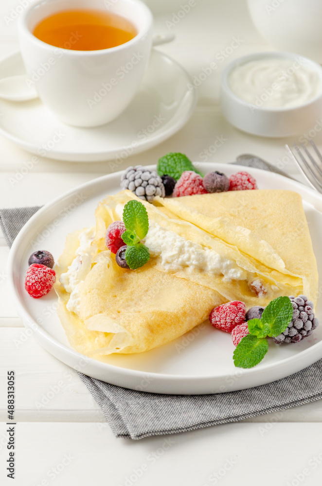 Traditional thin pancakes or crepes with cottage cheese and raisins with berries, honey and sour cream on a white wooden background. Food for Maslenitsa. Copy space.