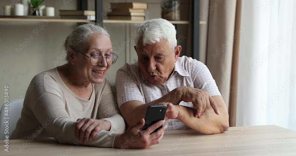 Smiling 80s retired couple, grey-haired wife and husband sit at table at home using cellphone, looking at gadget screen watch funny online videos, new blog, laughing enjoy modern tech amusing content Smiling 80s retired couple, grey-haired wife and husband sit at table at home using cellphone, looking at gadget screen watch funny online videos, new blog, laughing enjoy modern tech amusing content