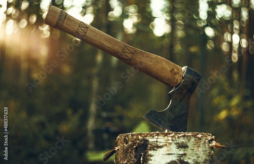 An axe with a wooden handle is stuck in a wooden log. Logging on the background of sunset. The hatchet. photo