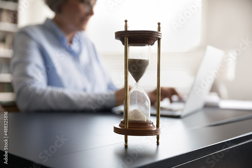 Close up focus on hourglass or sand clock standing on table with blurred middle aged senior businesswoman on background working on laptop, meeting deadline, time management, efficiency concept.