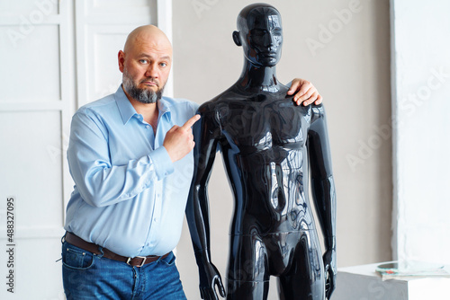 Bald sad adult man with beard, holding shoulders of naked artificial plastic black manikin, looking to dummy, thinking of alopecia treatment. No hair, replacement medicine concept. Light indoor photo