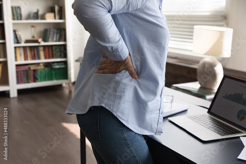 Close up cropped stressed mature old woman touching lower back, suffering from sudden pain after long sedentary work on computer, feeling physical discomfort overworking online at modern home office.