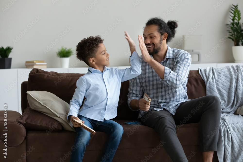 Happy little cute African American kid boy giving high five to caring millennial father, playing entertaining war game with wooden guns, enjoying domestic hobby activity pastime, having fun indoors. Happy little cute African American kid boy giving high five to caring millennial father, playing entertaining war game with wooden guns, enjoying domestic hobby activity pastime, having fun indoors.
