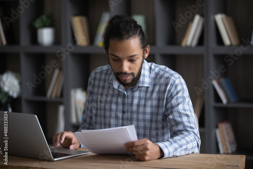 Focused smart 30s millennial African American male manager employee worker holding paper documents, analyzing economic report preparing presentation on computer, reviewing sales data statistics.