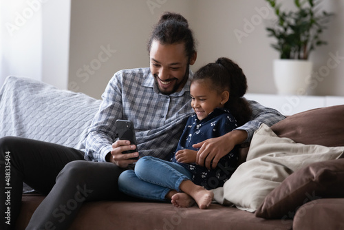 Cheerful affectionate 30s African American father showing funny videos online on cellphone to laughing adorable small kid daughter, enjoying playing games, posing for selfie photos, tech addiction. © fizkes