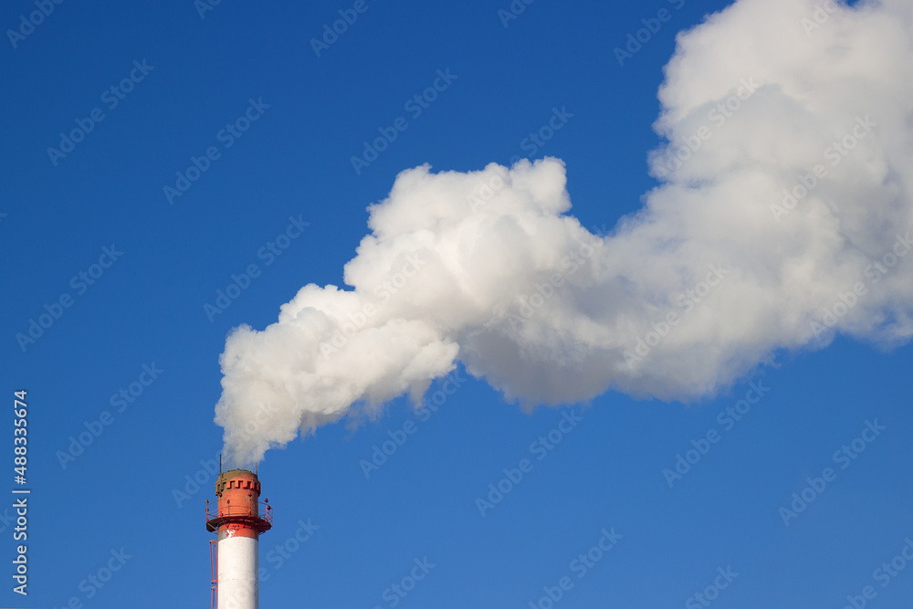 White thick smoke above a red and white pipe of boiler-house against blue sky. Winter heating season. Industrial background. Air pollution from fuel combustion