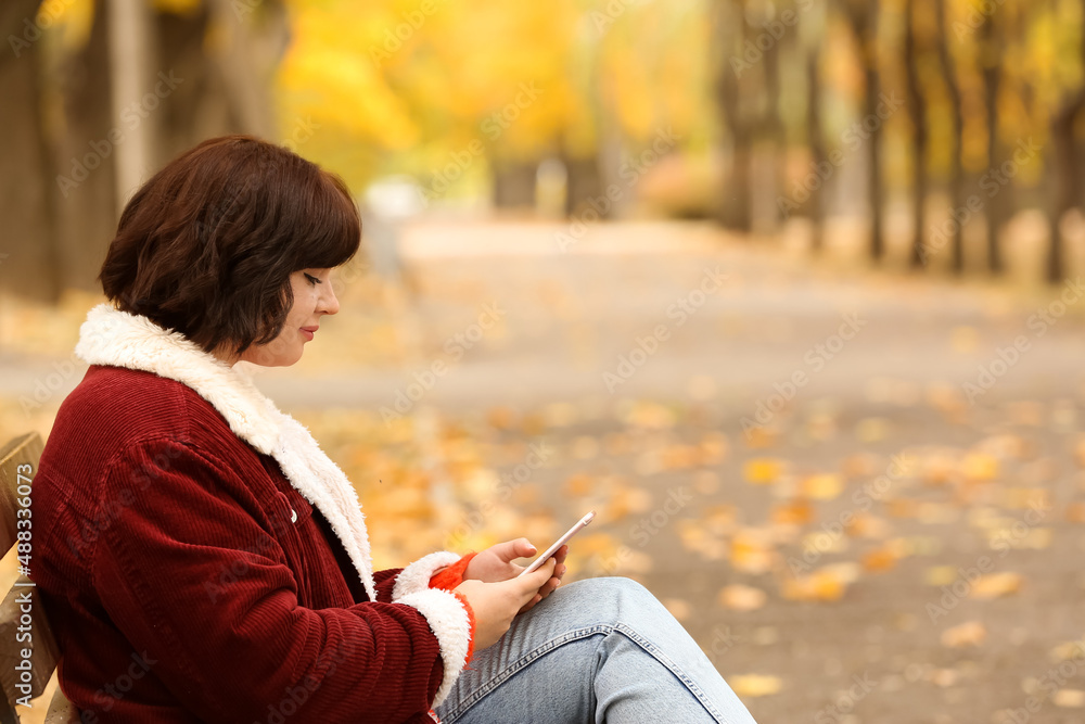 Beautiful brunette woman sitting on bench and using mobile phone in autumn park Beautiful brunette woman sitting on bench and using mobile phone in autumn park