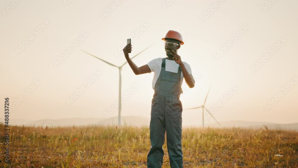 Positive engineer in uniform and helmet using modern smartphone for video call among windmill farm. Concept of people, technology and green energy. Working on eco station. Positive engineer in uniform and helmet using modern smartphone for video call among windmill farm. Concept of people, technology and green energy. Working on eco station.