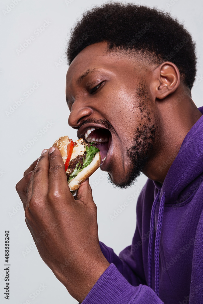 An African American man in a purple hoodie with short curly hair and beard open his mouth widely to bite a burger An African American man in a purple hoodie with short curly hair and beard open his mouth widely to bite a burger