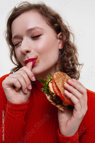 A vertical portrait of a curly girl a red turtleneck licking finger after eating cheeseburger with sauce eyes closed