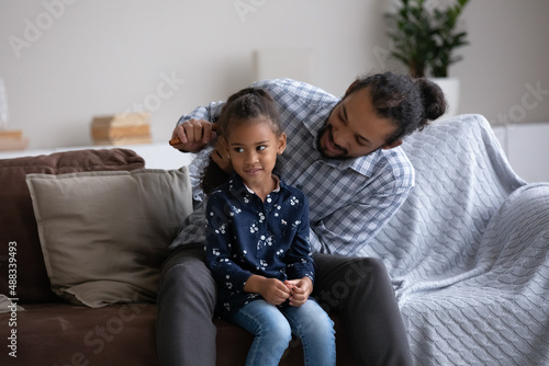 Smiling young African American father detangling long curly hair of cute happy little preteen child daughter, making hairstyle helping getting ready for school, sitting together on couch at home. photo