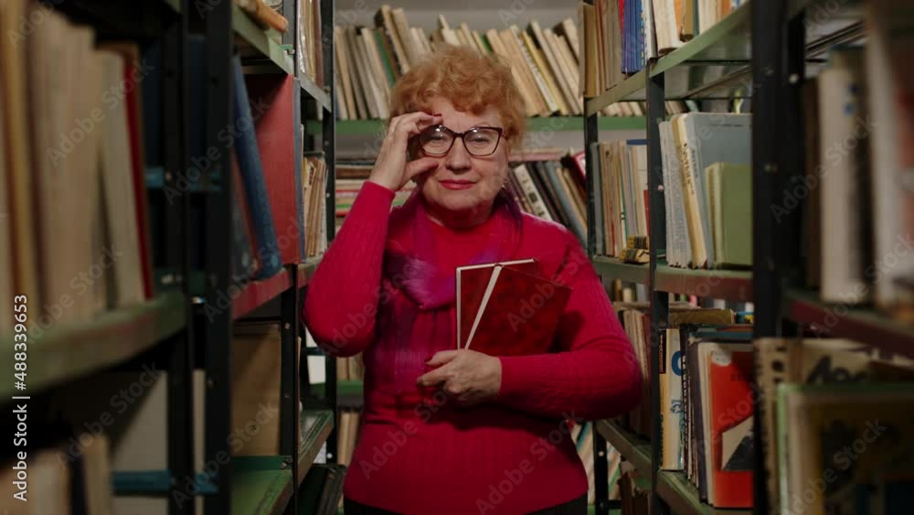 Smart elderly teacher in glasses holding books in her hands in the library Smart elderly teacher in glasses holding books in her hands in the library