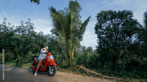 Woman on red motorbike in white clothes drive on forest road trail trip. One girl caucasian tourist go on scooter, nearby tropical palm tree. Asia Thailand ride tourism. Motorcycle rent Safety helmet.