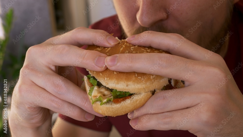 Close-up of a bearded man eating an appetizing burger, a juicy cutlet with vegetables and sauces in a sesame seed bun. Vegetarian hamburger with meat substitute.